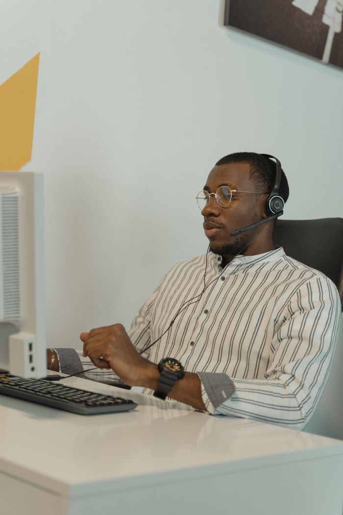 Black man wearing glasses and headset working at a computer in an office setting.